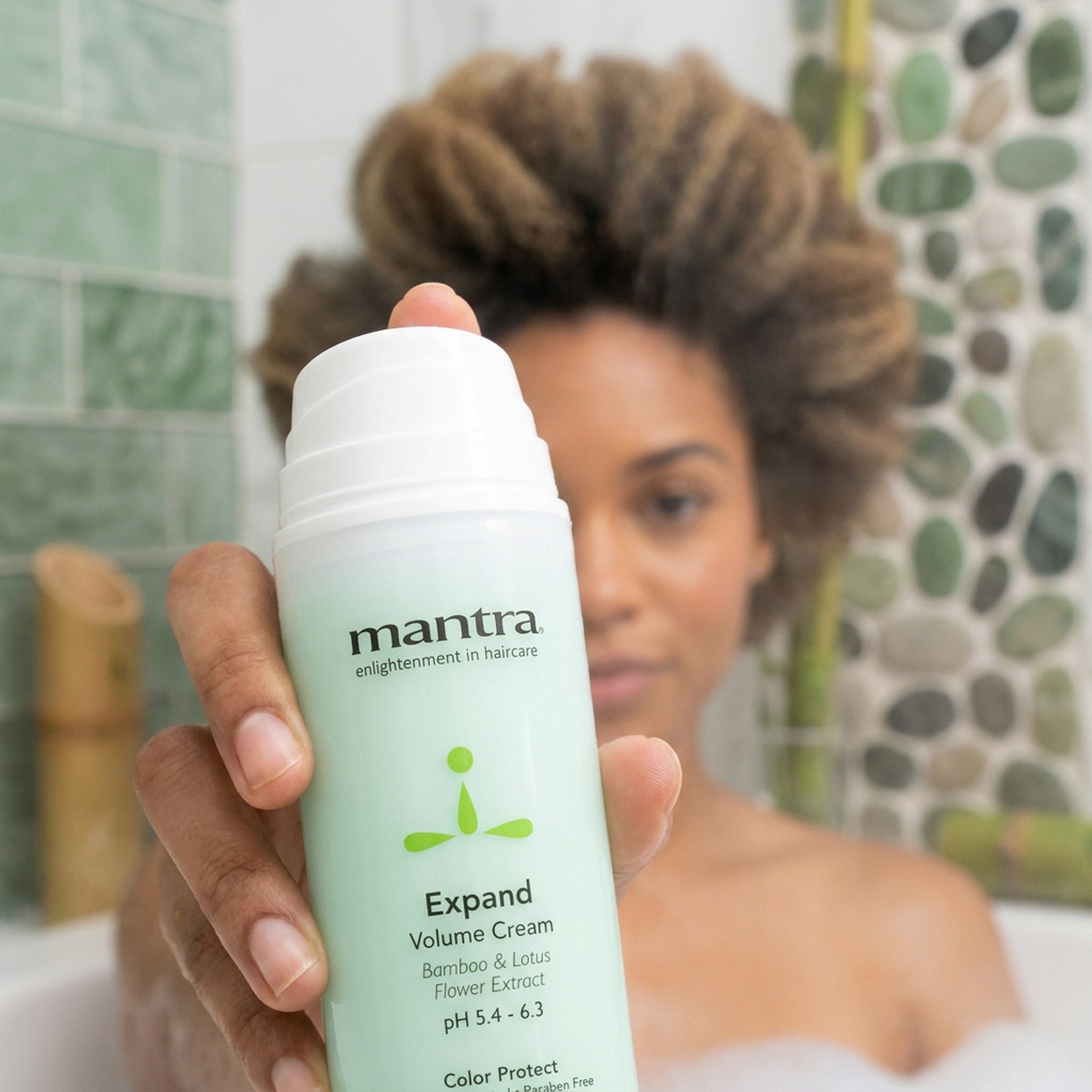 Woman holding a bottle of Mantra Expand Volume Cream up to the camera in a soft green bathroom