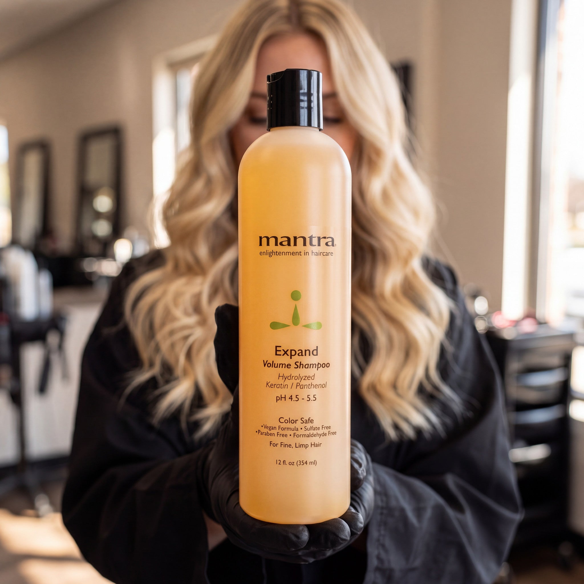 Stylist in a black salon smock holding a bottle of Mantra Expand Volume Shampoo up to the camera in a salon
