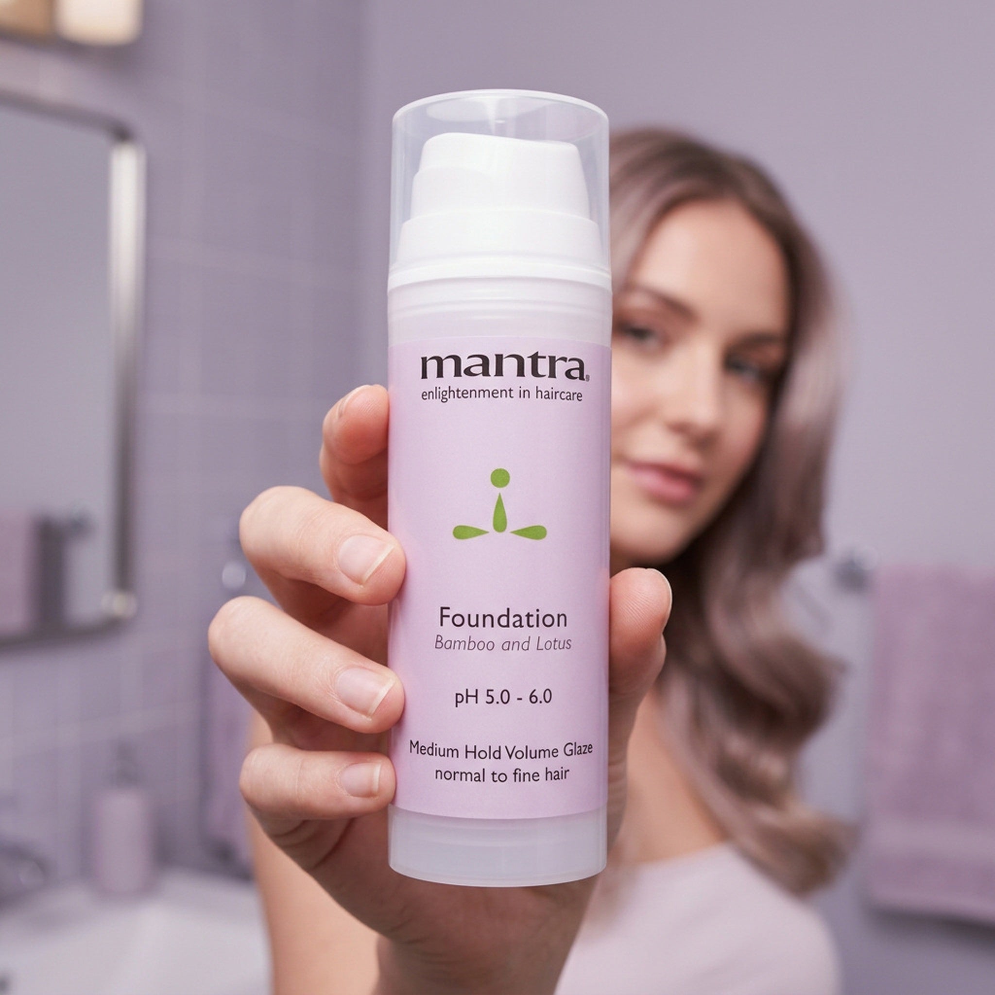 Woman holding a bottle of Mantra Foundation Volume Glaze up to the camera in a lilac bathroom