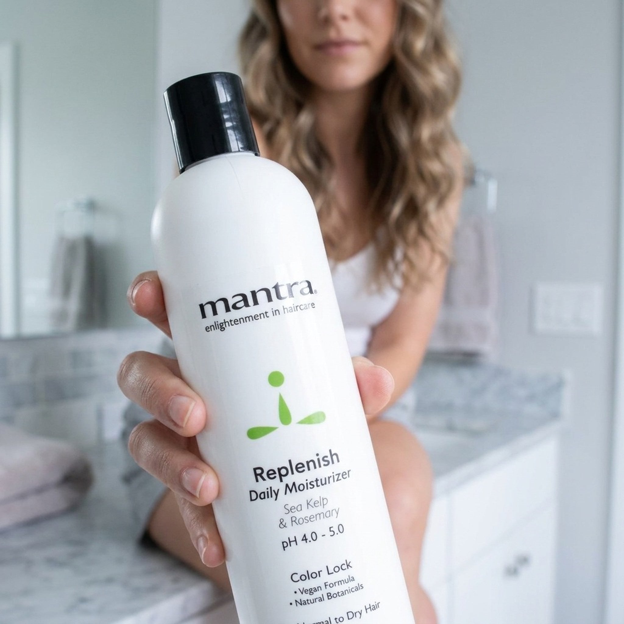 Woman holding a bottle of Mantra Replenish Daily Conditioner up to the camera in a soft white bathroom