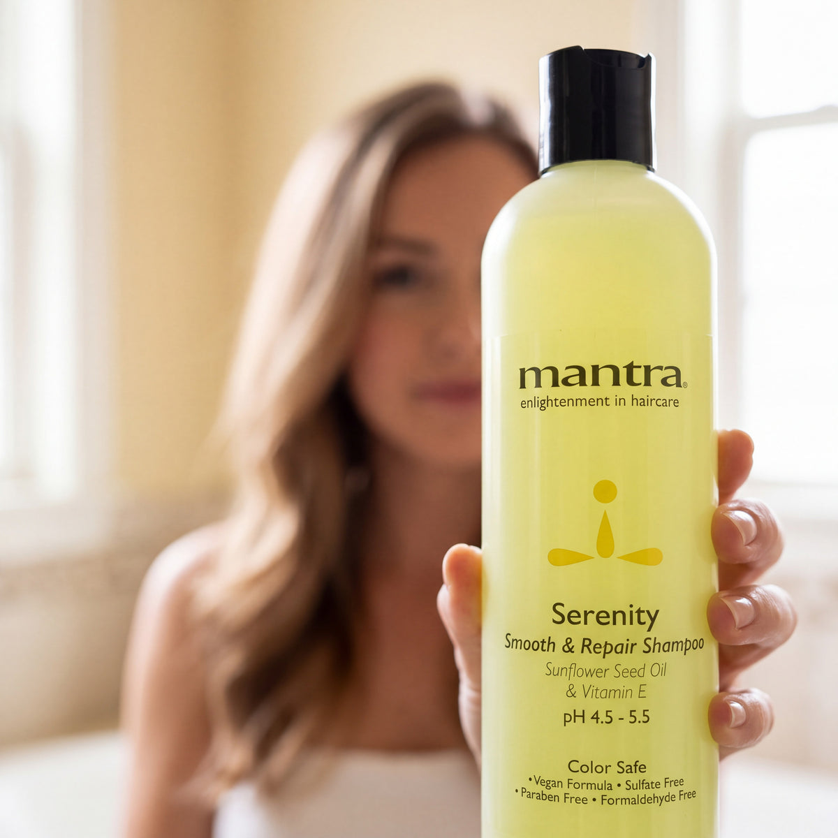 Woman holding a bottle of Mantra Serenity Daily Shampoo up to the camera in a warm neutral bathroom