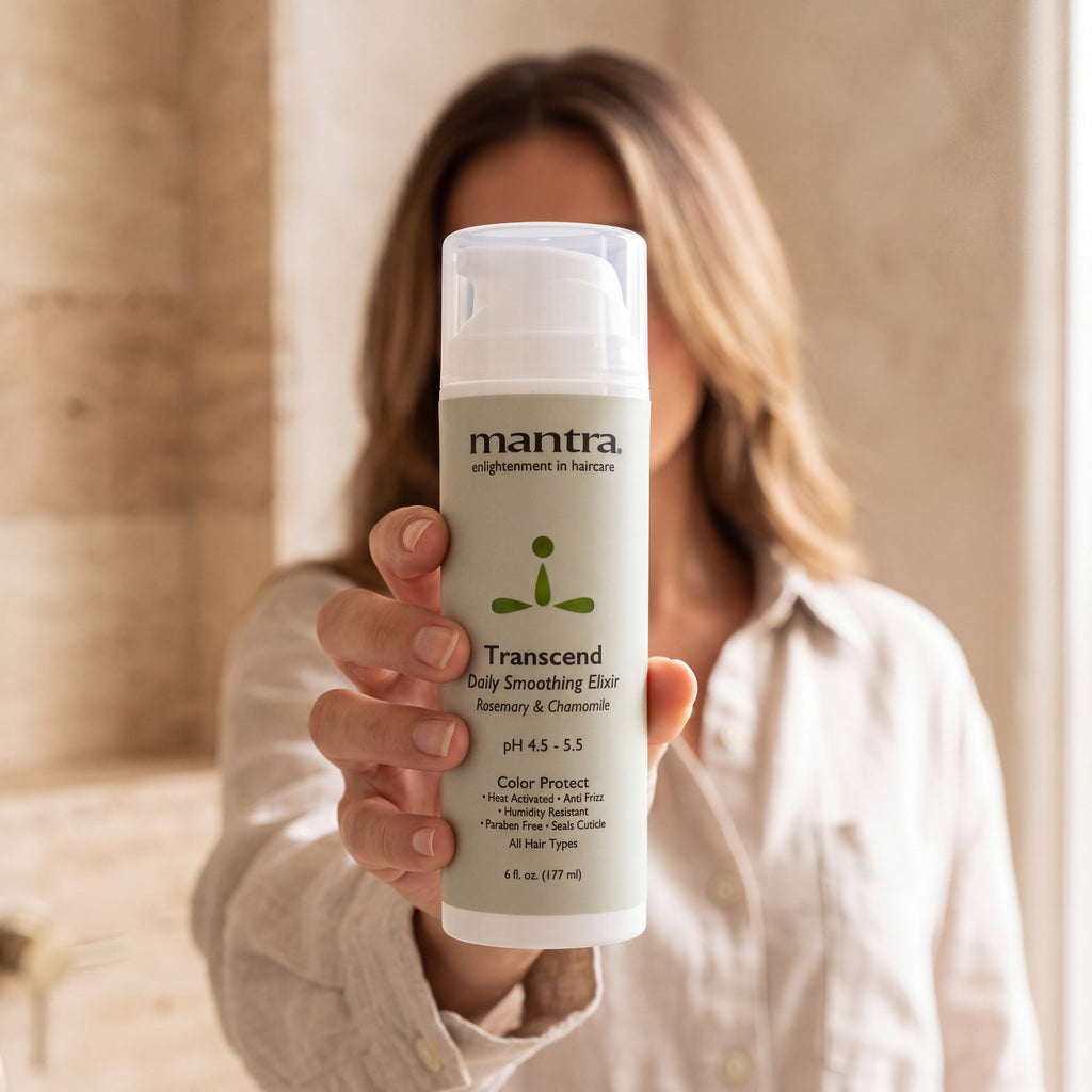 Woman in an oatmeal linen shirt holding a bottle of Mantra Transcend Daily Smoothing Elixir up to the camera in a sunlit bathroom